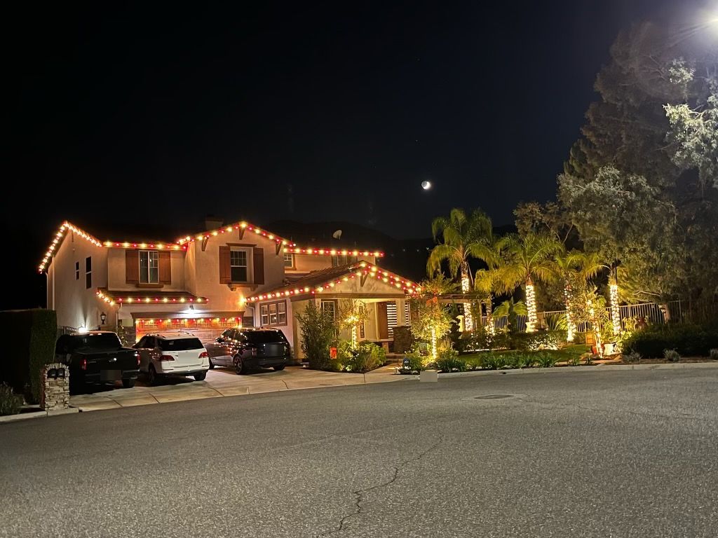 Two-story estate with red and warm-white roofline, a warm-white-wrapped palm courtyard, and a crescent moon overhead